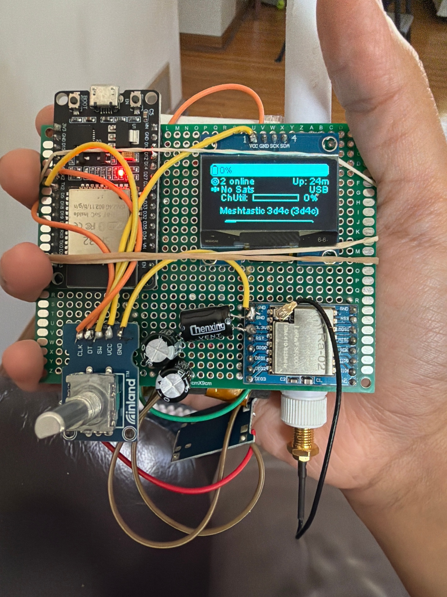 A handheld Meshtastic LoRa device built on perfboard with an ESP32, OLED display, rotary encoder, and SMA antenna.